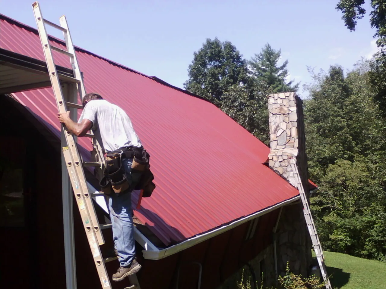 Roofer on a ladder beside a red metal roof and stone chimney — Soffit & Fascia Repair by Roofing Brothers in Ironton